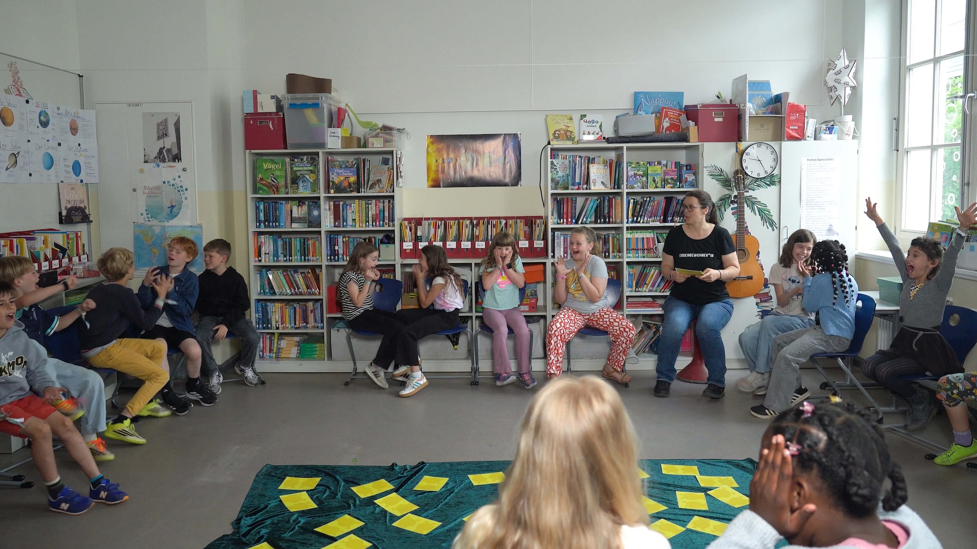 Klasse im Sitzkreis in der Schulbibliothek, einige Kinder melden sich